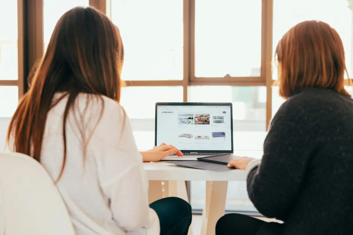 Dos personas de espaldas revisando una página web en una laptop junto a una ventana luminosa
